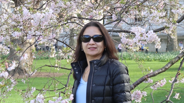 Nidhi Bansal, a woman with long brown hair wearing sunglasses, a black puff jacket and a light blue t-shirt, stands in front of blooming cherry blossom trees.