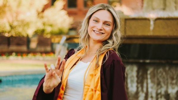 Paige Oskolkoff sitting on the edge of a water fountain wearing her graduation robes and stole, making the ASU pitchfork sign with her hands and smiling.