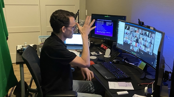 ASL lecturer Paul Quinn, wearing light blue jeans and a black polo shirt, sits in a black chair in front of a desk covered with several computer monitors. The faces of his students are visible on one screen. He is demonstrating a sign with his right hand.