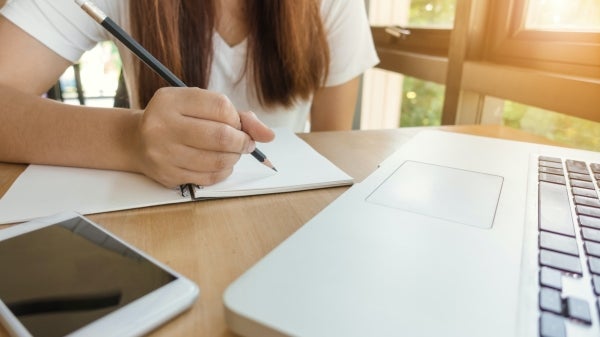 Person writing in notebook with phone and computer on desk in front of her.