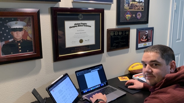 ASU student Ryan Johnston seated at a desk with a laptop. The wall behind the desk features photos and memorabilia from his time in the military.