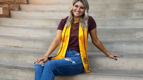 Sarah Bruder sits on concrete steps, smiling at the camera with her gold graduation sash and maroon cords draped around her neck