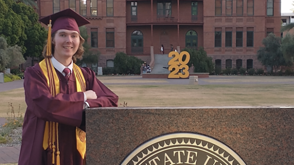 A student in a graduation cap and gown poses next to an ASU sign
