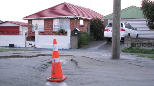 Photo showing liquefaction in a neighborhood setting with houses and cars around and an orange cone on a damaged street.