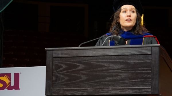 Woman standing behind a lectern wearing graduation regalia and speaking into a micorphone.