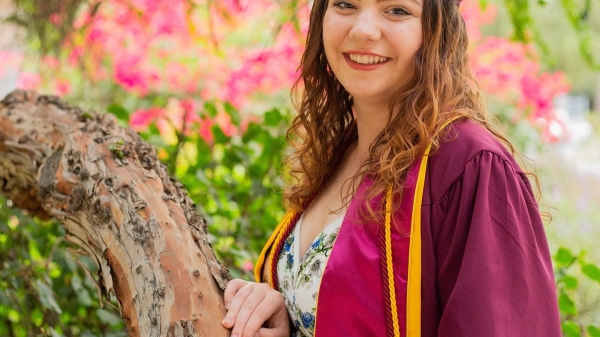 Sydney Campton poses in her maroon graduation robe over a light-colored top. Curly light brown hair hangs down from under her maroon graduation cap. Her hand is resting on a tree branch and there are out of focus leaves and flowers behind her.