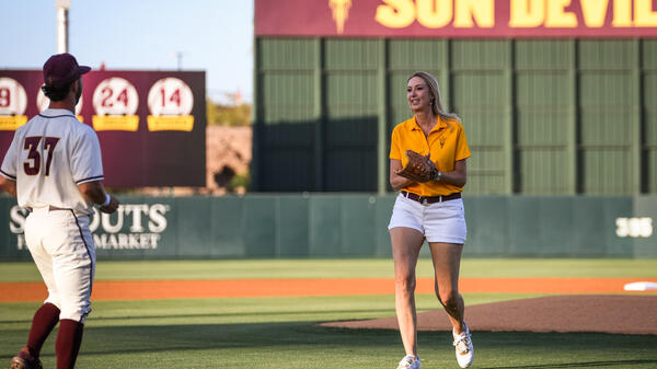 Woman wearing a baseball mitt on a baseball field, walking toward a baseball player in uniform.