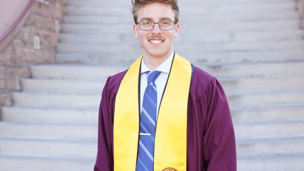 Student wearing academic regalia stands outside at the bottom of the staircase at Old Main.