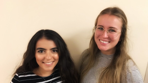 Jasmine Dahdal and Zoe Zimmerman smile at the camera. Jasmine has a dark striped shirt on and Zoe has a light striped shirt on. They are standing against a beige background