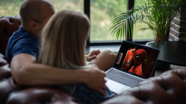 A couple seen from behind, seated on a couch while streaming a movie on a laptop.