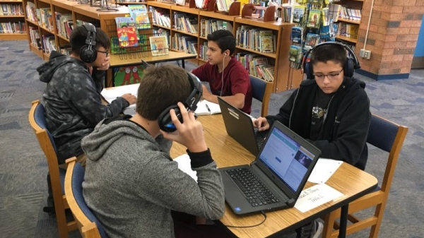 Four middle school students sit at a table in a school library logged in to a digital class, wearing headphones in front of laptops