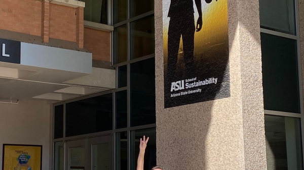 A man stands outside holding his hand in a pitchfork gesture, mimicking an ASU marketing sign above him.