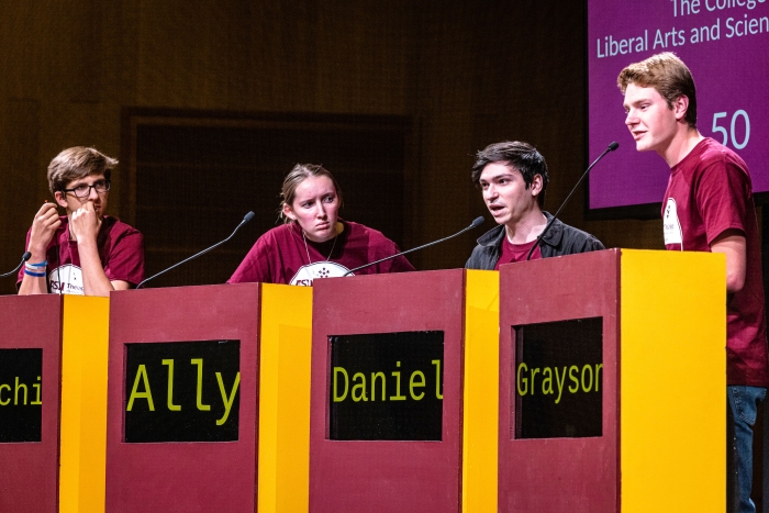 Four members of a quiz bowl team stand behind lecterns at a competition