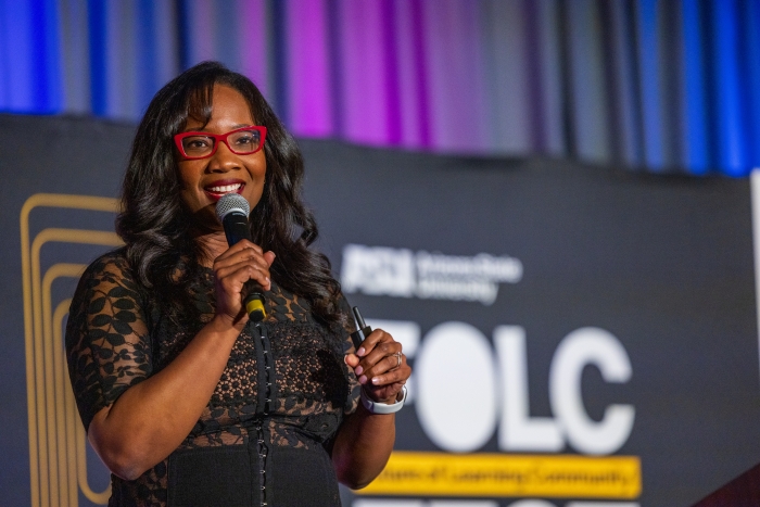 Black woman with long hair wearing red glasses speaks into microphone on stage