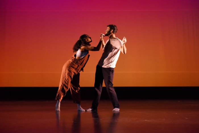 One dancer points while another moves away as they perform against an orange background