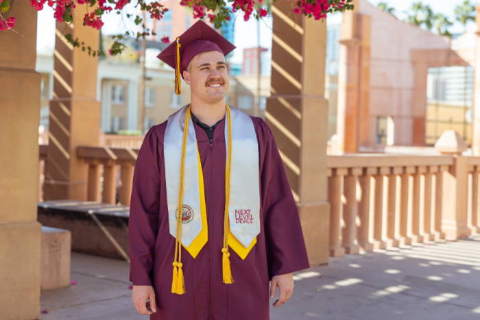 An ASU graduate in commencement cap and gown smiles off camera while standing near Hayden Library
