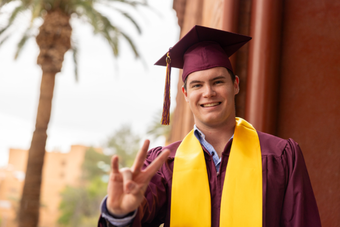 An ASU graduate smiles at the camera while holding up a pitchfork gesture