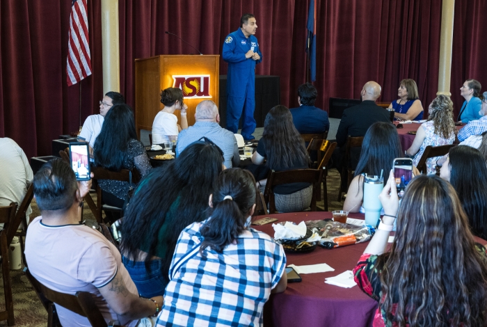 Man wearing a NASA flight suit speaking to a crowded room.