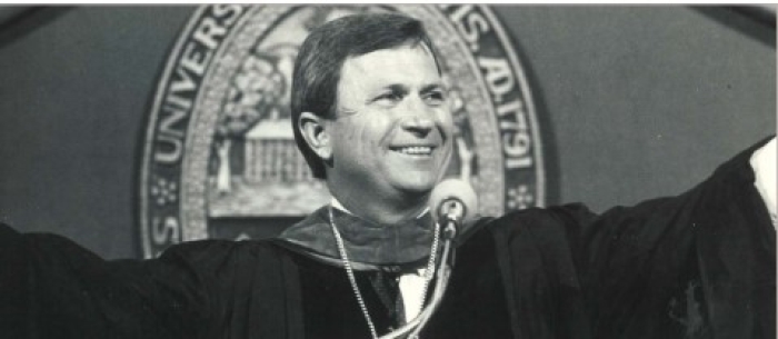 A man in graduation regalia holds his arms wide