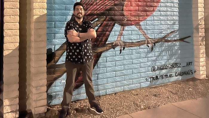 Man standing with crossed arms in front of a mural on a brick wall.