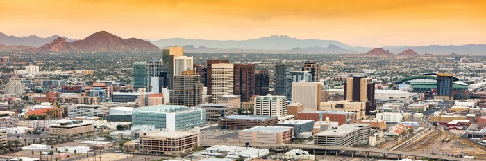 Panoramic view of Phoenix's skyline at dusk with skyscrapers, mid-rise buildings, and mountain ranges in the background under an orange sky.