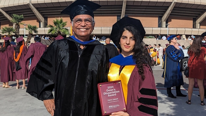 A student wearing graduation regalia holds a plaque while standing next to a faculty member.