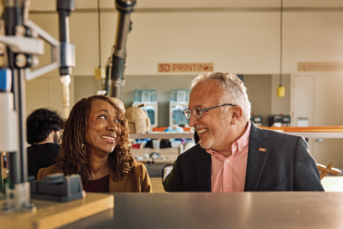 Photo shows a Black woman with medium length thin locs and white man with gray hair and classes smiling at each other in a Makerspace workshop