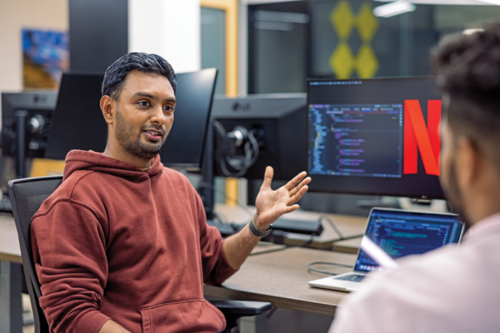 Young man in a brick red hoodie talks to a colleague at a desk of computers