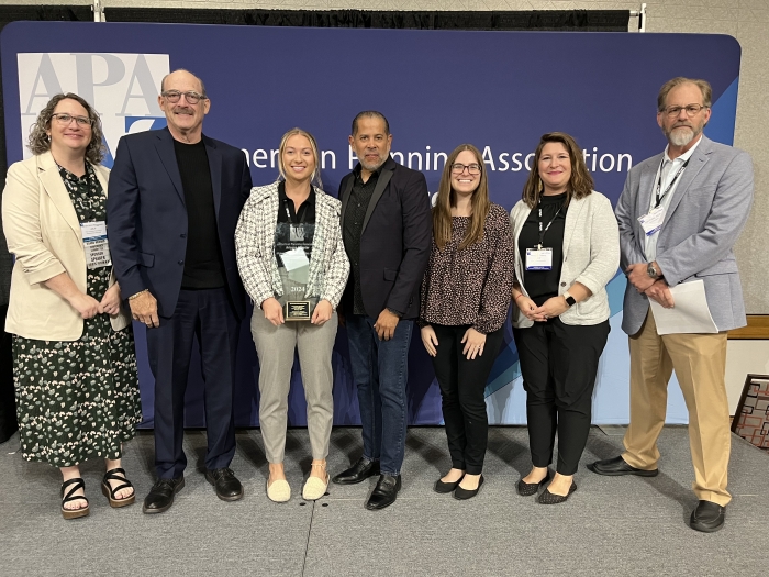 Seven people stand in a line for a photo with one person holding an award