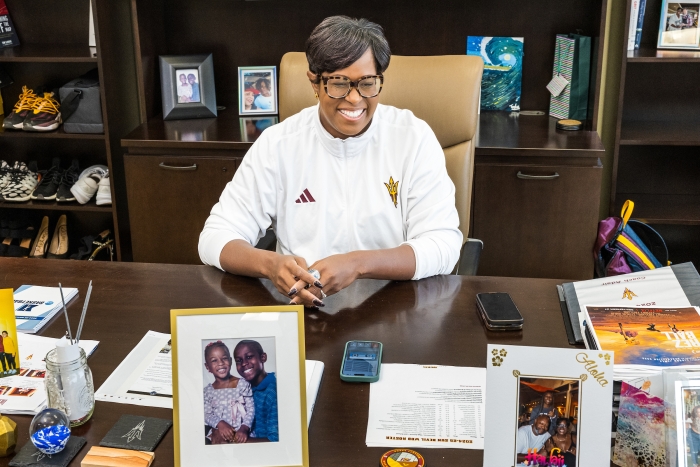 Black woman with short hair wearing glasses and a white tracksuit jacket sits behind a desk filled with pictures of family