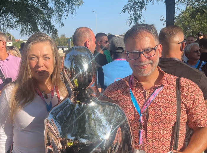 Man and woman standing next to chrome monument in the shape of a human.