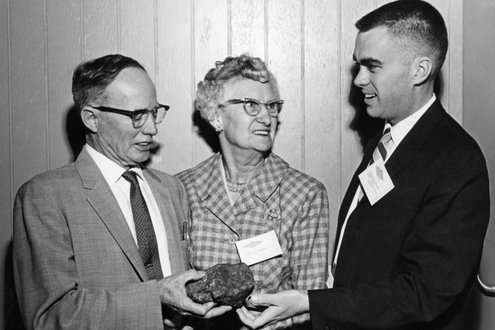 Black and white photo of an older man in a suit handing over a meteorite to a younger man in a suit while an older woman in the middle looks on