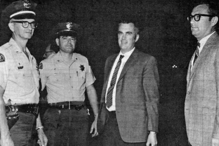 Black and white photo of two police officers escorting two men in suits