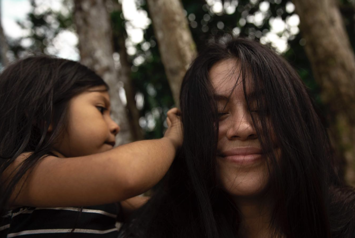 A child looking through Elizabeth's hair, both with dark hair, smiling softly outdoors with blurred trees in the background.