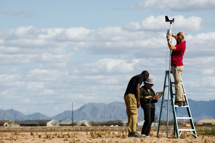 Scott stands on a ladder to install equipment on a pole above a barren field while two researchers study laptop