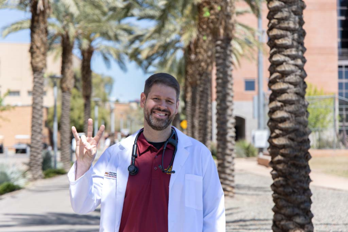 Chris Stone smiles at the camera. He is wearing a white nurse practitioner coat with a black stethoscope around his neck.