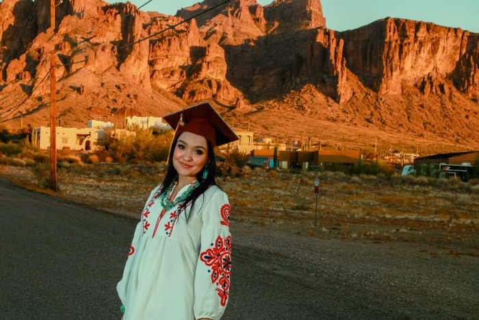 Esperanza Biggs smiles at the camera wearing her ASU graduation cap and a white shirt with red flowers on the sleeves. A mountain range can be seen in the background