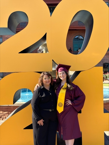 A mom and her daughter in cap and gown pose for a photo in front of a giant 2024 sign