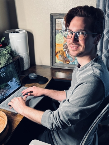 Justin Brengel sits at his desk while typing on his laptop and turns to smile at the camera