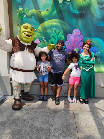 Man and his two children stand with Shrek and Princess Fiona at a theme park.