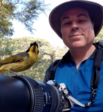 A man poses for a photo holding a camera in front of him, on which sits a bird