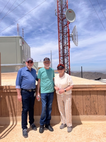 Two men and a woman stand in front of a mountaintop broadcast tower.