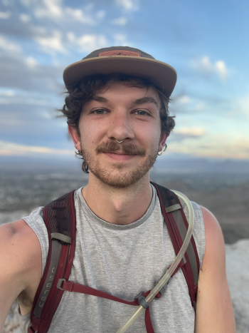 A man with a mustache wearing a backpack smiles at the camera from a mountaintop.