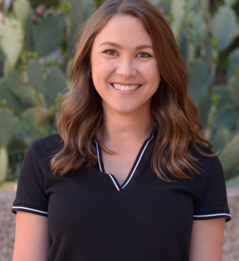 A woman in a black shirt with brown hair smiles at the camera in front of a prickly pear cactus.