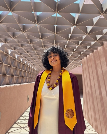 Danielle Ochoa, wearing her ASU graduation robes and a white dress, smiles at the camera while posing under a geometric walkway. 