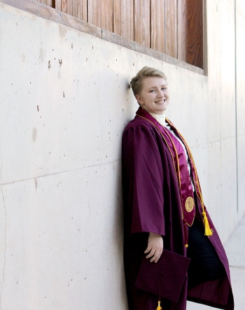 A graduate in regalia leans against a wall