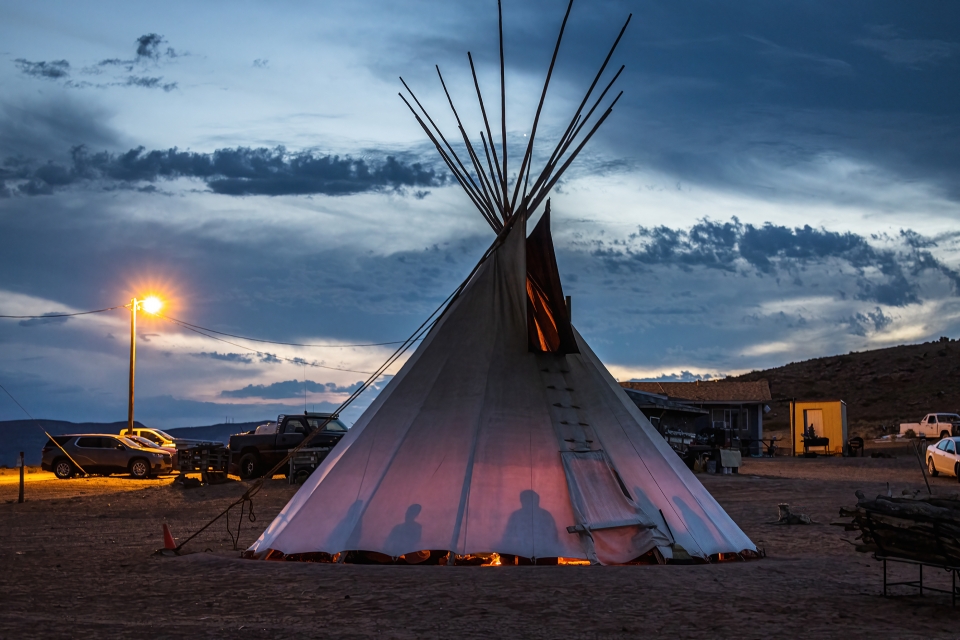 A teepee lit from a fire inside is shown against a sunset sky; several people are silhouetted inside