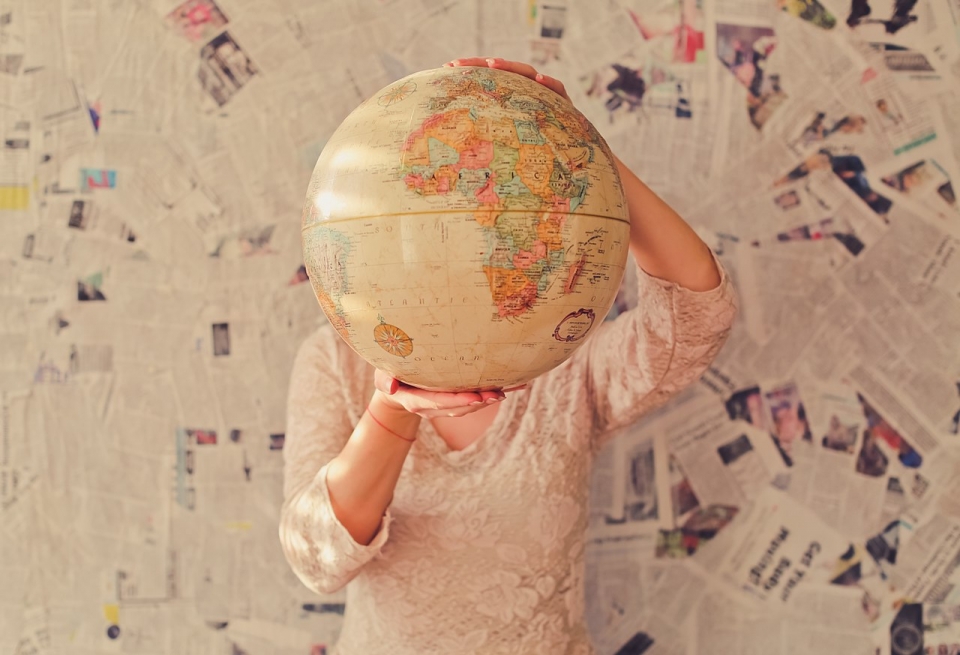 A person holds a globe in front of a wall of newspapers. / Public domain image credit: Slava Bowman on Unsplash, via Wikimedia Commons.
