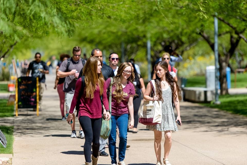 Groups of students walk down a wide sidewalk surrounded by desert trees on the Polytechnic campus
