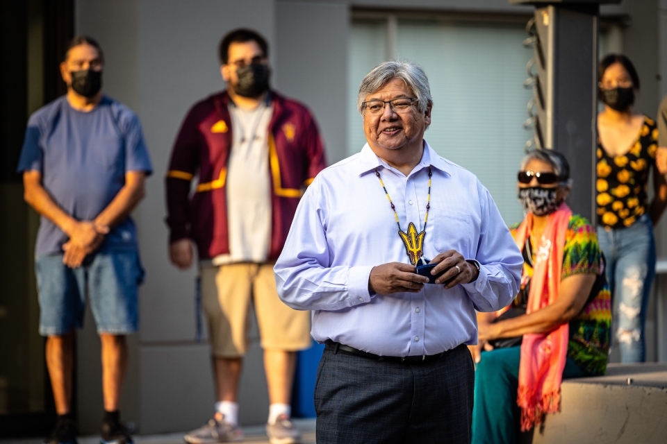 A Native American man wearing a beaded ASU pitchfork medallion speaks outside at dawn to a group